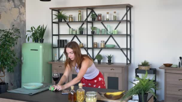Krásná mladá hospodyně stírací tabulku s ručníkem a zvedl sklenku vína v kuchyni během dopoledne, koncentrované a stanoví — Stock video