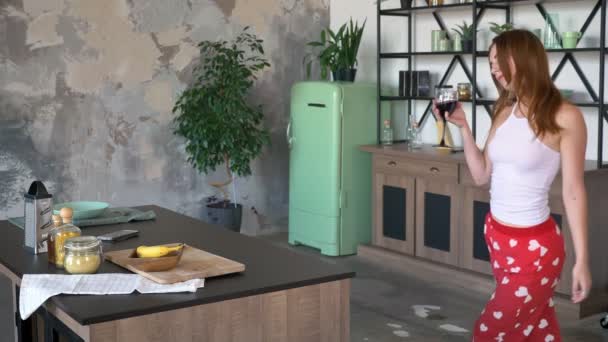 Preciosa joven con cabello de jengibre bailando y sosteniendo copa de vino, cocina moderna con mesa grande y nevera en el fondo — Vídeos de Stock