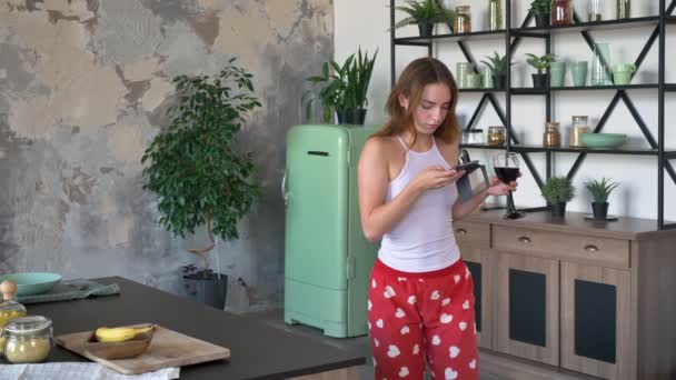 Atractiva mujer seria con cabello de jengibre sosteniendo vino de vidrio y charlando por teléfono, bailando en cocina con mesa grande y nevera — Vídeo de stock