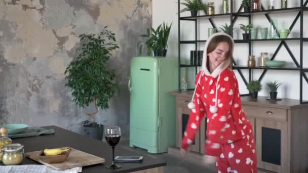 Happy young woman in pajamas dancing and smiling, wearing fancy hood, cheerful and funny, modern kitchen with green fridge — Stock Video