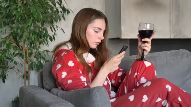 Charming ginger woman typing on phone and holding glass of wine, sitting on couch in modern living room, serious and pensive — Stock Video