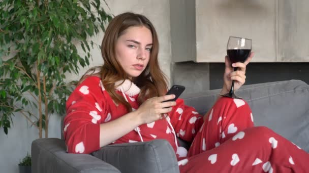 Fundersam ung kvinna hålla glas vin och att skriva på telefonen, sittande på soffan i modernt vardagsrum — Stockvideo