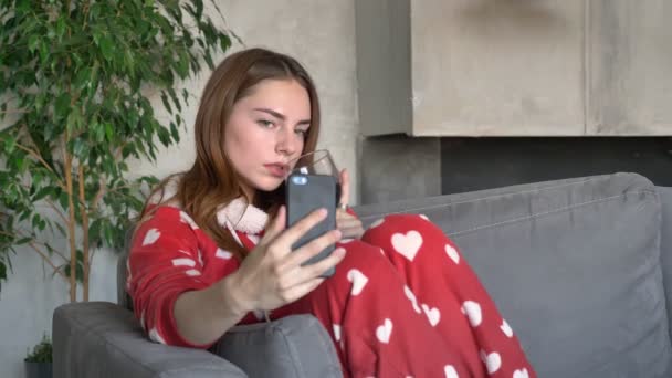 Belle femme rousse prenant selfie avec un verre de vin, assise sur le canapé dans le salon moderne, portant un pyjama rouge dans les cœurs — Video