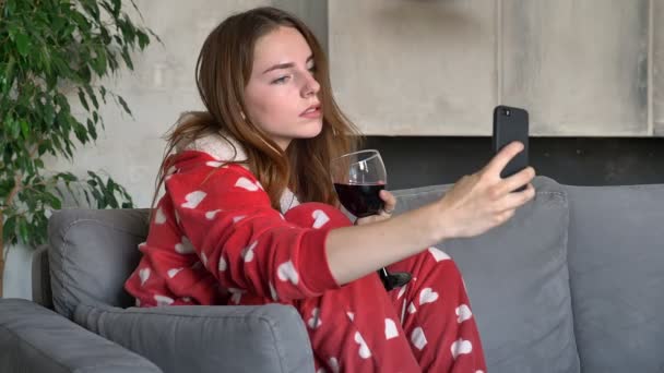 Jovem encantadora posando com copo de vinho e tomando selfie, sentada no sofá na sala de estar, vestindo pijamas engraçados com corações — Vídeo de Stock