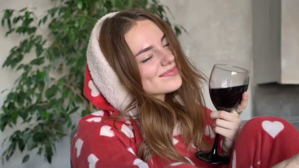 Cheerful ginger woman wearing hood of pajamas and taking selfie, holding glass of wine and sitting on couch in living room — Stock Video