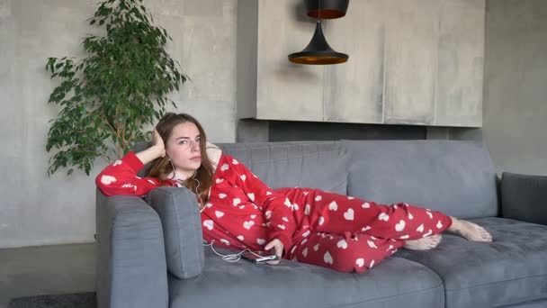 Красивая рыжая женщина в красной пижаме лежит на диване и слушает музыку через наушники в гостиной днем — стоковое видео