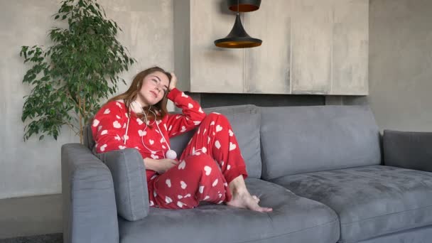 Encantadora joven sentada en el sofá y escuchando música a través de auriculares, teléfono, sala de estar moderna en el fondo — Vídeos de Stock