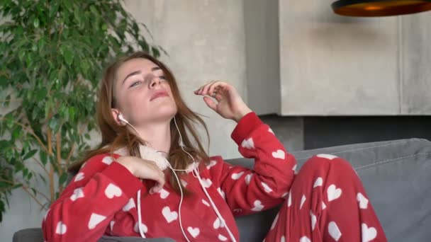 Привлекательная молодая женщина в красной пижаме танцует и слушает музыку через наушники, сидит на диване в гостиной, днем — стоковое видео