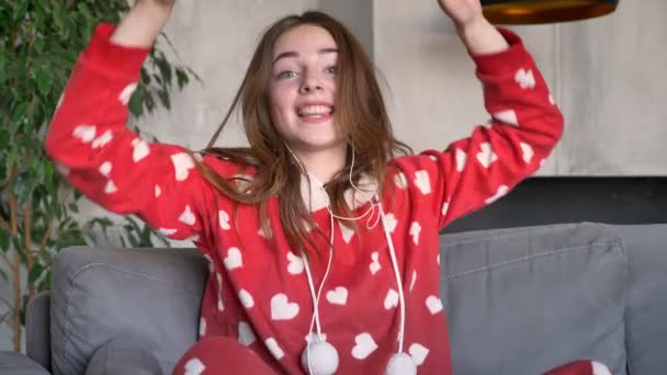 Glada ingefära kvinna sitter på soffan och dans när du lyssnar musik genom hörlurar, modernt vardagsrum i bakgrunden — Stockvideo