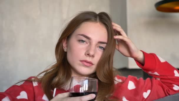 Portrait of beautiful woman with ginger hair looking at camera and holding glass of wine, sitting on couch in modern living room — Stock Video