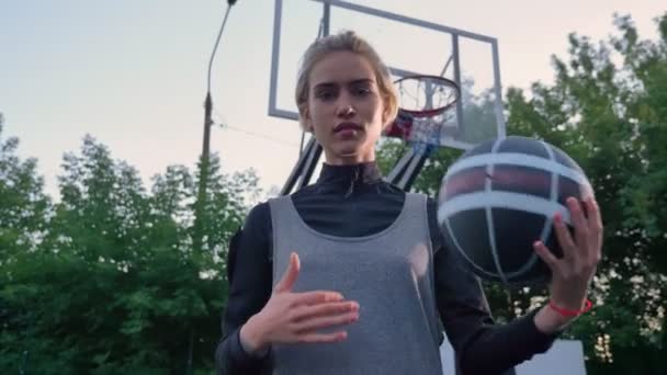 Pemain bola basket wanita pirang yang menarik bermain dengan bola dan melihat ke kamera, berdiri di taman, siang hari — Stok Video