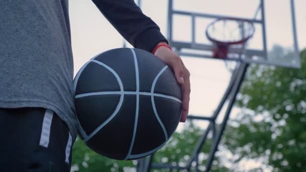 Близкие кадры женской руки, держащей баскетбол, стоящей в парке, обруч на заднем плане, днем — стоковое видео