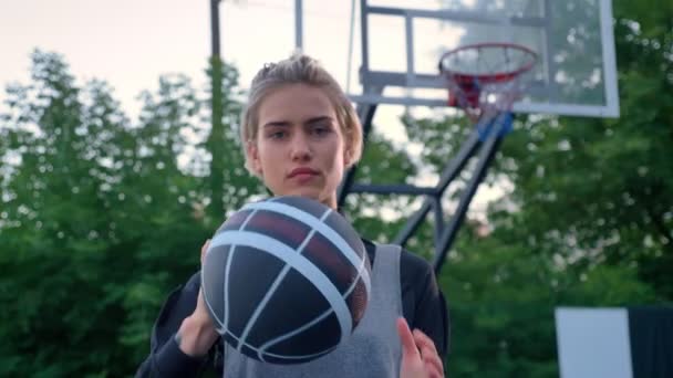 Krásná blondýnka ukazující basketbal do kamery, profesionální hráč stojící v parku během dne, obruč v pozadí — Stock video