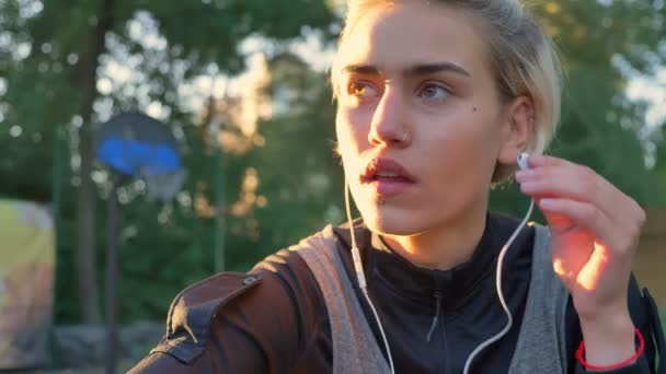 Beautiful blonde woman with pierced nose wearing earphones, sitting in park during sunset, looking forward — Stock Video