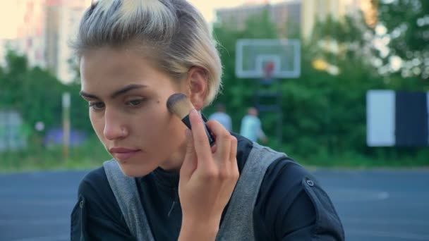 Sluiten de beelden van prachtige vrouwelijke basketballer toepassing van make up en zittend op de grond, mannen spelen op achtergrond — Stockvideo