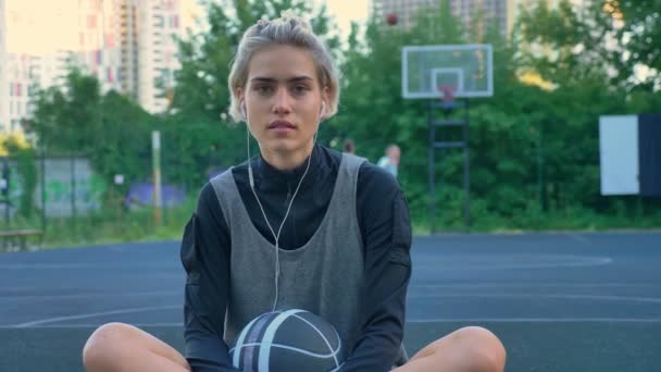 Sorrindo jovem jogador de basquete feminino ouvindo música através de fones de ouvido e sentado na quadra, homens jogando no fundo no parque — Vídeo de Stock