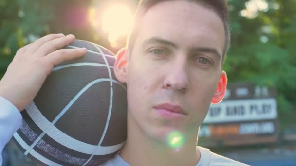 Porträt eines jungen selbstbewussten Mannes, der Basketball hält und in die Kamera blickt, im Hintergrund parkt — Stockvideo