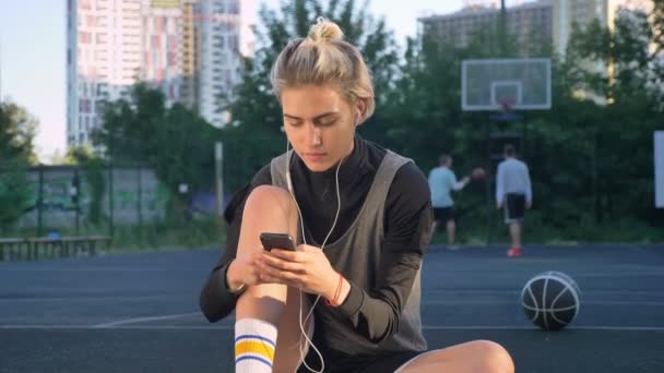 Pensive young beautiful woman sitting on basketball court and typing on phone, wearing earphones, men playing in background — Stock Video