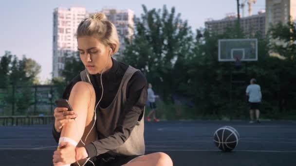 Hermosa mujer rubia escuchando música a través de auriculares y escribiendo en el teléfono, sentado en la cancha, hombre en el fondo lanzando pelota — Vídeos de Stock