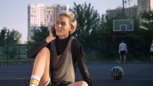 Gelukkig vrouwelijke basketballer praten over telefoon en glimlachend, zittend op de Hof in open lucht, mannen in het spelen van de achtergrond — Stockvideo