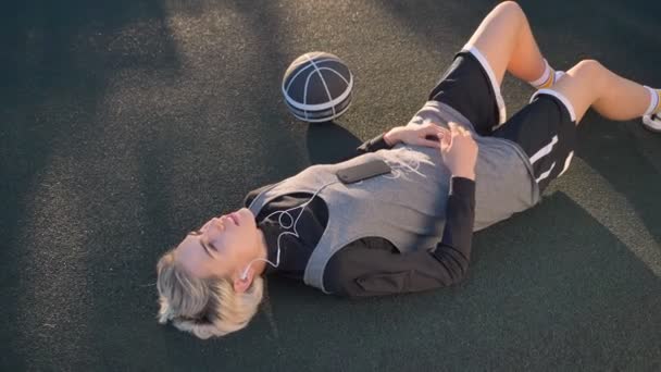 Баскетболистка-блондинка лежит на корте и слушает музыку, женщина отдыхает на свежем воздухе во время заката — стоковое видео
