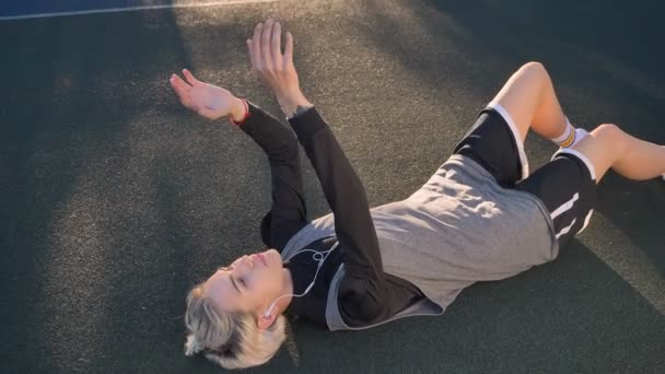 Encantadora mujer rubia tumbada en el suelo y jugando al baloncesto, escuchando música a través de auriculares, hermosa jugadora femenina — Vídeo de stock