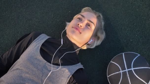 Drömmande unga kvinnliga basketspelare liggande på domstolen och lyssna på musik genom hörlurar, bollen att placera nära henne, fridfullt och lugnt — Stockvideo