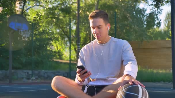 Красивый счастливый молодой человек сидит на баскетбольной площадке и печатает по телефону, держит мяч и слушает музыку, улыбается — стоковое видео