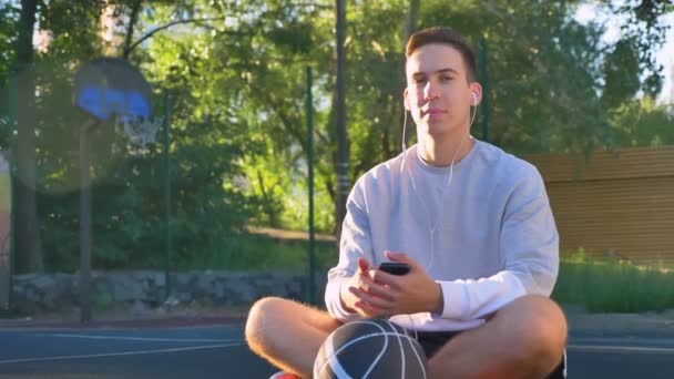 Giovane fiducioso seduto sul campo da basket e ascoltare musica, guardando la fotocamera, tenendo il telefono e la palla, parco in background — Video Stock