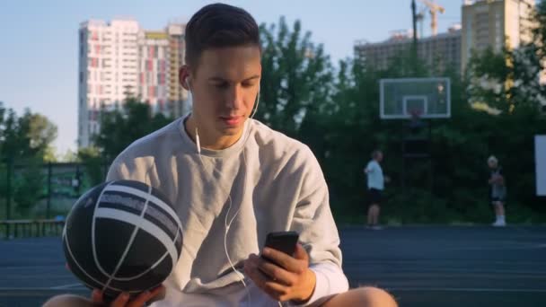 Joven guapo sentado en la cancha de baloncesto y escribiendo en el teléfono, mirando a la cámara, sosteniendo la pelota, la gente jugando en el fondo — Vídeo de stock