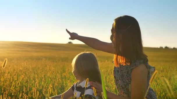 Joven madre sentada en el campo con la pequeña hija rubia y señalando al atardecer, mujer feliz con chica al aire libre, sonriendo — Vídeos de Stock