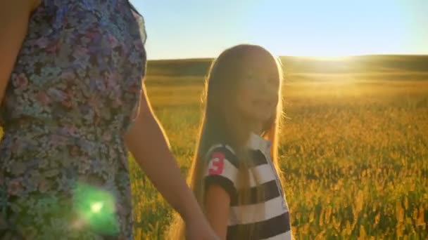 Hermosa niña rubia caminando en el campo de trigo y mirando a la madre, cogida de la mano y sonriendo, apuntando a la puesta de sol en el fondo — Vídeos de Stock