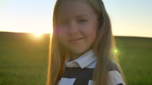 Retrato de hermosa niña con el pelo rubio sonriendo a la cámara, campo con puesta de sol en el fondo, niño feliz y alegre — Vídeos de Stock