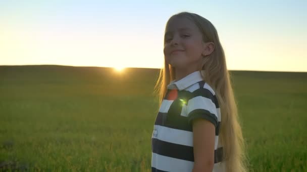 Hermosa niña rubia de pie en el campo de trigo y sonriendo a la cámara, hermosa vista durante la puesta de sol en el fondo, niño feliz — Vídeos de Stock