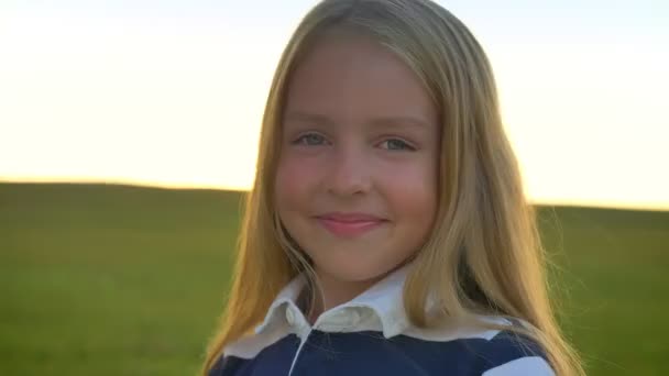Portret dziewczynka Blondynka o niebieskich oczach, uśmiecha się do kamery, stojąc na białym polu pszenicy i żyta podczas zachodu słońca w tle — Wideo stockowe