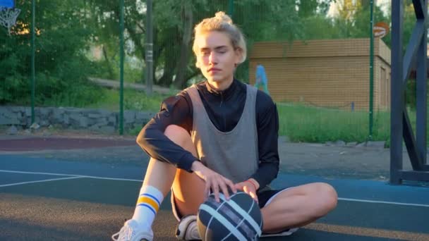 Mooie blonde vrouwelijke basketballer zittend op de grond en spelen met de bal, park in achtergrond — Stockvideo