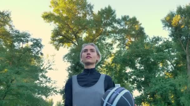 Charmante joueuse de basket-ball marchant et jouant avec la balle, parc avec ciel bleu au-dessus en arrière-plan, angle bas — Video