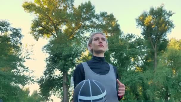 Уверенная блондинка-баскетболистка держит мяч и ходит в парке, под низким углом, носит спортивную одежду — стоковое видео