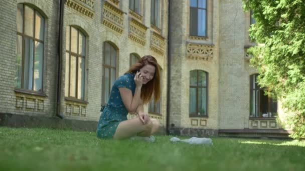 Young ginger woman talking on phone and sitting on grass in park with building in background, beautiful sunny day outdoors — Stock Video