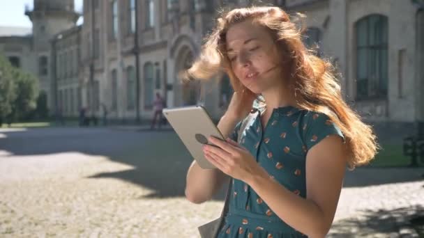 Hermosa joven jengibre escribiendo en la tableta, viento volando su pelo, de pie en la calle y sonriente, feliz y alegre hembra — Vídeos de Stock