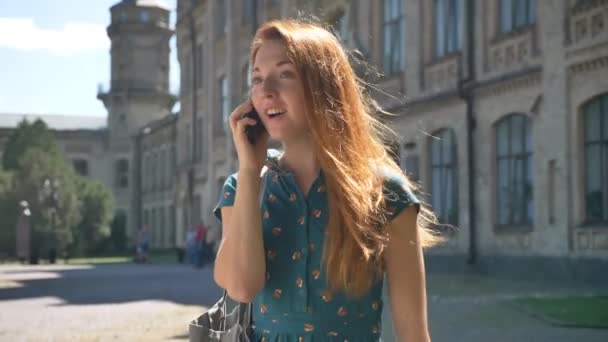 Šťastná mladá zázvor žena mluví o telefonu a s úsměvem, stojící na ulici, budovu v pozadí, vítr ve vlasech — Stock video