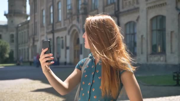 Kız öğrenci gülümseyen görüntülü sohbet sahip ve arka planda bina ile sokakta duran mutlu zencefil genç kadın — Stok video