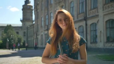 Genç güzel şık güneş gözlüğü kameraya, seyir gülümseyerek ve düşünme, arka planda Bina kadında zencefil