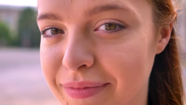 Retrato cercano de una joven mujer pelirroja tímida mirando a la cámara, calle en el fondo, hermosa mujer sonriendo al aire libre — Vídeo de stock