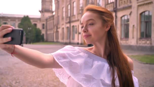 Beautiful ginger young woman with bare shoulders taking selfie with her phone and smiling, standing on street with building in background — Stock Video