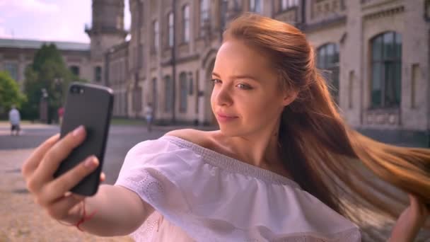 Adorable young ginger woman with bare shoulders taking selfie and smiling, touching her hair, standing on street — Stock Video