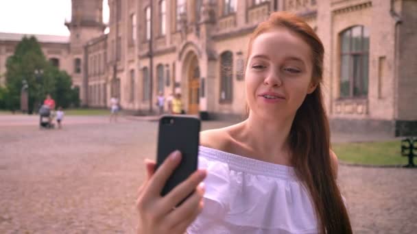 Beautiful young ginger woman with bare shoulders having video chat and standing on street with building in background, smiling — Stock Video