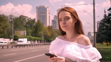 Telefon tutarak ve taksi, yol yakınında sokakta gündüz ayakta bekleyen çıplak omuz olan dalgın zencefil kadın