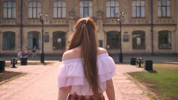 Вид сзади на студентку с длинными рыжими волосами, идущую в университетское здание в парке, в рубашке с голыми плечами — стоковое видео