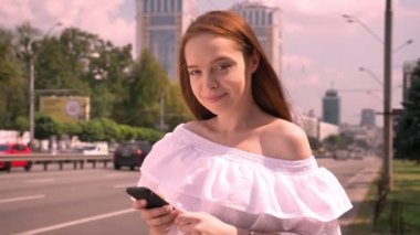 Güneşli gün boyunca yol yakınında sokakta taksi bekliyor telefon tutarak ve kameraya, seyir çekici genç zencefil kadın